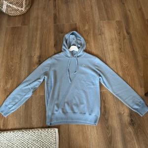 Ljusblå hoodie från Bvaldi - Snygg ljusblå hoodie från Bvaldi med huva och snörning. Tröjan är stickad i mjukt material och har ribbade muddar vid ärmslut och nederkant. Perfekt för dig som gillar en clean och enkel stil. Helt ny, köpt för 7 dagar sedan, var lite liten på mig. Köpt för 1200.