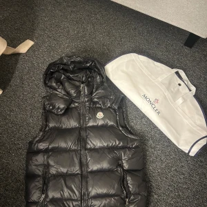 Svart dunväst från Moncler - Svart dunväst från Moncler med glansig finish, avtagbar huva och dragkedja framtill. Västen har två fickor med dragkedja och klassisk Moncler-logga på bröstet. Perfekt för lager-på-lager och snygg streetstil.