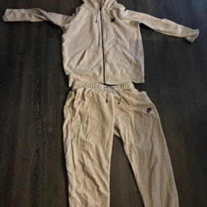Beige hoodie från Nike med dragkedja med ett par begie mjukis  - Säljer en beige sätt från Nike med dragkedja framtill och huva med snörning. Tröjan har långa ärmar och två fickor på magen. Perfekt för chill dagar eller när du vill vara extra bekväm.