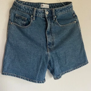 Blå denimshorts Zara - Klassiska blå denimshorts från Zara med hög midja och fem fickor. Raka ben och snyggt avslut vid benslutet. Perfekta för sommaren och enkla att matcha med allt. Stängs med knapp och dragkedja framtill.