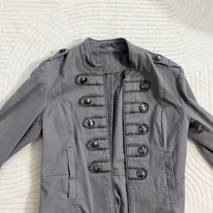 Grå rock med militärstil och knappar - Snygg grå rock med militärinspirerad design, dekorerad med svarta knappar och broderade detaljer framtill. Jackan har ståkrage, långa ärmar och dragkedja. Perfekt för dig som gillar en cool och unik look.