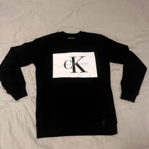 Svart sweatshirt från Calvin Klein - Svart sweatshirt från Calvin Klein Jeans med stor vit logga på bröstet. Tröjan har rund hals, långa ärmar och ribbade muddar vid ärmslut och nederkant. Perfekt för dig som gillar stilrena och klassiska plagg med tydlig branding.