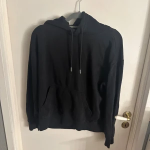 Svart hoodie från H&M med huva - Svart hoodie från H&M i storlek S. Klassisk modell med huva och snörning, samt en stor magficka framtill. Mjuk och skön bomullsmix, perfekt för chill dagar. Långärmad och loose fit för en avslappnad vibe.
