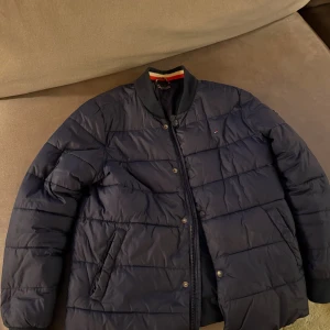 Mörkblå pufferjacka Tommy Hilfiger - Snygg mörkblå pufferjacka från Tommy Hilfiger med tekniska fibrer. Jackan har ribbad krage, dragkedja och tryckknappar framtill samt två sidofickor. Perfekt för kyliga dagar och har en stilren design med diskret logga på bröstet. Storlek 164