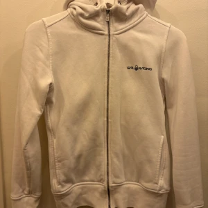 Vit hoodie från Sail Racing XS - Vit hoodie från Sail Racing i storlek XS. Tröjan har dragkedja framtill, huva och två fickor. Broderad logga på bröstet och på huvan. Mjuk insida och klassisk passform, perfekt för en clean och sportig look.