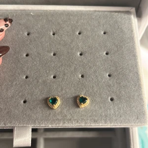 Guldörhängen med gröna hjärtan - Säljer ett par små örhängen i guldton med hjärtformad grön sten i mitten och glittriga detaljer runt om. Nypris 300 