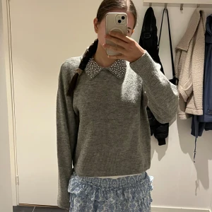 Grå stickad tröja med pärlkrage - Superfin grå stickad tröja med lång ärm och en snygg krage dekorerad med pärlor. Perfekt för dig som vill ha något stilrent men ändå med en twist. Tröjan har en klassisk passform och ribbade muddar vid ärmslut och nederkant.