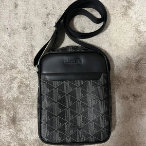 Svart axelväska från Lacoste med mönster - Snygg svart axelväska från Lacoste med geometriskt mönster i grått och silver. Väskan har justerbar axelrem med metallspänne och präglad Lacoste-logga på framsidan. Perfekt storlek för mobil, plånbok och andra småsaker.