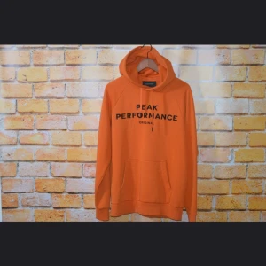 Orange hoodie från Peak Performance - Snygg orange hoodie från Peak Performance med svart broderad logga på bröstet. Tröjan har huva med dragsko, känguruficka och långa ärmar. Perfekt för dig som gillar streetwear och vill sticka ut med en färgstark look.