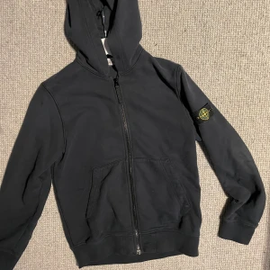 Stone Island Hoodie junior men passar xs - Svart hoodie från Stone Island med klassisk logga på ärmen. Tröjan har dragkedja framtill, huva och två stora fickor. Tillverkad i mjuk bomull, perfekt för en chill och stilren look. Knappt använd, pris kan diskuteras. Köpt på farfetch