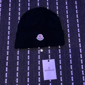 Svart ribbad mössa från Moncler - Svart ribbstickad mössa från Moncler med klassisk uppvikt kant och logotyp framtill. Perfekt för att hålla värmen och samtidigt se clean ut. Snygg och enkel design som funkar till alla jackor. Materialet är mjukt och bekvämt.