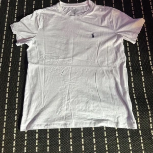 Vit t-shirt från Polo Ralph Lauren - Vit t-shirt från Polo Ralph Lauren med klassisk rund hals och liten broderad logga på bröstet. Modellen är relaxed fit och tillverkad i mjuk bomull som känns skön mot huden. Perfekt för dig som gillar stilrena och enkla plagg.