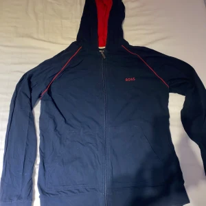 Marinblå hoodie från BOSS med röd huva - Snygg marinblå hoodie från BOSS med hel dragkedja och röda detaljer. Insidan av huvan är röd och tröjan har diskret BOSS-logga på bröstet. Tillverkad i mjuk bomull och har långa ärmar samt fickor framtill. Perfekt för en chill och stilren look.