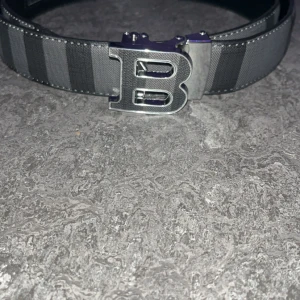 Randigt svart bälte med B-spänne - Snyggt svart bälte med breda, mörka ränder och ett stort metallspänne i form av bokstaven B. Bältet har en stilren design med tydliga sömmar och är gjort i ett tåligt material med läderlook. Perfekt accessoar för att lyfta din outfit.