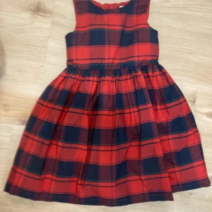 Rutig röd klänning Holly & Whyte - Snygg rutig klänning från Holly & Whyte by Lindex i storlek 116. Klänningen är ärmlös med rund hals och har ett klassiskt rött och mörkblått rutmönster. Kjolen är utställd och midjan markerad, perfekt för dig som gillar färg och mönster. Köparen betalt för frakten.
