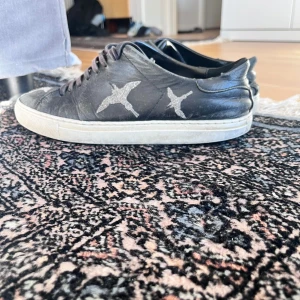 Svarta sneakers med fågeltryck - Svarta sneakers i skinn med vita fågeltryck på sidorna. Skorna har snörning, platt vit sula och rund tå. Snygg kontrast mellan det svarta lädret och de vita detaljerna. Perfekta för dig som gillar unika detaljer på dina skor.