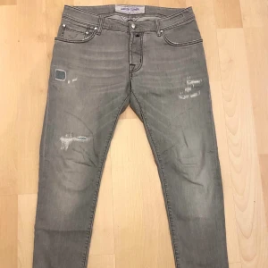 Grå jeans från Jacob Cohën - Snygga grå jeans från Jacob Cohën i storlek 33. Modellen har klassisk femficksdesign, slitna detaljer och patchade partier framtill. Jeansen är tillverkade i bomull med lite stretch för skön passform och har raka ben med smal siluett. Mycket gott skick, inga defekter. Modell 622 W33
