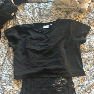 Svart croppad t-shirt & spetslinne Gina Tricot  - Två svarta toppar från Gina Tricot. Ena är en croppad t-shirt i bomull med kort ärm och v-ringning, andra är ett ärmlöst linne i genombruten spets med blommigt mönster. Perfekta att styla lager på lager eller var för sig.