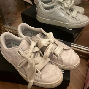 Vita sneakers från Puma med breda band - Säljer ett par vita Puma sneakers med breda, sidenliknande snören som ger en unik och trendig look. Skorna har klassisk rund tå, platt sula och subtila detaljer i skinnimitation. Perfekta för dig som vill ha en sportig men ändå söt stil.