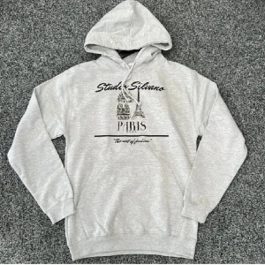 Grå hoodie Studio Silvano Paris - Grå hoodie från Studio Silvano med tryck av Paris och Eiffeltornet på bröstet. Tröjan har huva med snörning, magficka och ribbade muddar. Perfekt för dig som gillar streetstyle och vill ha en chill look. Materialet är mjuk bomull och polyester.