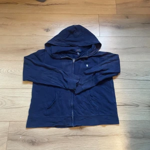 Marinblå hoodie från Polo Ralph Lauren - Snygg marinblå hoodie från Polo Ralph Lauren med dragkedja och huva. Tröjan har långa ärmar, två fickor fram och den klassiska Polo-loggan broderad på bröstet. Tillverkad i mjuk 100% bomull för skön komfort. Perfekt för chill dagar och streetwear.