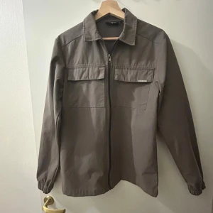 Brun overshirt med dragkedja - Snygg brun overshirt från Lindbergh med dragkedja framtill och två stora bröstfickor. Jackan har klassisk krage och långa ärmar med resår vid ärmslut. Perfekt för lager-på-lager och enkel att matcha med jeans eller chinos.