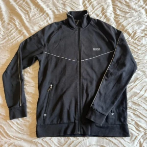 Svart ziptröja från Hugo Boss  - Svart långärmad ziptröja från BOSS med vit piping längs bröstet och ärmarna. Tröjan har ståkrage, två fickor med dragkedja och diskret BOSS-logga på vänster bröst. Perfekt för en chill och sportig look.