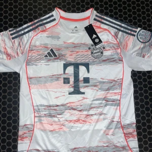 FC Bayern München bortatröja Adidas M - Snygg FC Bayern München bortatröja från Adidas i vitt med grå och röda grafiska detaljer. Tröjan har korta ärmar, rund hals och klassiska Adidas-ränder på axlarna. Tillverkad i lätt och ventilerande polyester med klubbmärke och sponsorlogga på bröstet. Bara höra av er vid frågor/funderingar Fraktar samma dag som köp🚚