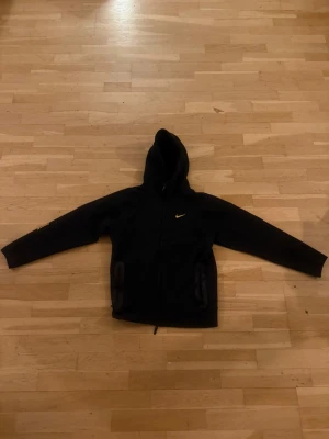 Svart Nike nocta zip hoodie med gul logga - Svart hoodie från Nike med dragkedja och huva. Klassisk gul Nike Swoosh på bröstet och två fickor framtill. Tillverkad i mjukt material som känns skönt mot huden. Perfekt för en sportig och avslappnad stil.