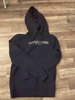 Mörkblå hoodie från Jack & Jones - Snygg mörkblå hoodie från Jack & Jones med vit broderad logga på bröstet. Klassisk modell med huva och långa ärmar. Perfekt för en avslappnad och trendig stil. Storleken är xs/s, den är i riktigt fint skick.