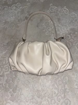Krämvit scrunchie handväska - Trendig krämvit handväska i scrunchie-modell med mjukt, rynkat syntetmaterial. Väskan har ett avtagbart handtag och guldfärgade metalldetaljer. Stängs med dragkedja och har svart innerfoder. Perfekt accessoar för att lyfta din outfit.