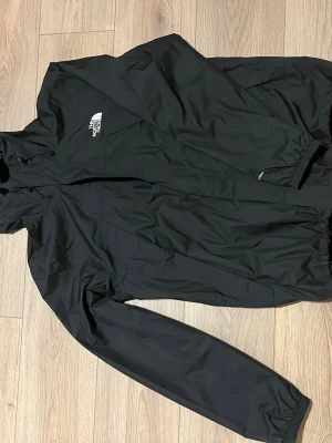 Svart vindjacka från The North Face - Helt ny snygg svart vindjacka från The North Face med huva och dragkedja framtill. Jackan har logga på bröstet och ryggen, elastiska muddar vid ärmslut och nederkant samt en klassisk, normal passform. Perfekt för blåsiga dagar. Köpte för 1500kr men den är fortfarande helt ny men den var för stor för mig. 