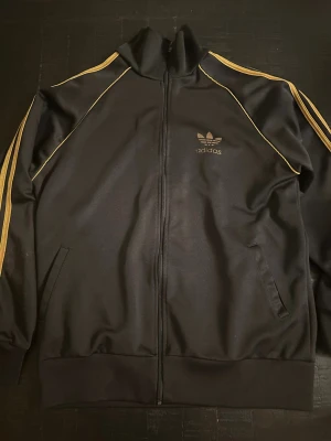 Svart Adidas track jacket med guld - Snygg svart track jacket från Adidas med guldfärgade detaljer och klassiska ränder längs ärmarna. Dragkedja framtill, hög krage och fickor på sidorna. Adidas logga på bröstet och stor logga på ryggen. Perfekt för en sportig och retro look.