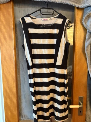 Randig ärmlös klänning Indiska - Snygg ärmlös klänning från Indiska med breda svarta och vita horisontella ränder. Klänningen är i kort modell och har en rund halsringning. Tillverkad i mjukt bomullsmaterial som känns skönt mot huden. Perfekt för en avslappnad och trendig look.