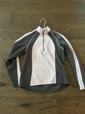 Grå och vit träningströja från Craft - Säljer en långärmad träningströja från Craft i grått och vitt. Tröjan har halv dragkedja framtill, ståkrage och är tillverkad i ett ribbat, tekniskt material som andas. Perfekt för löpning eller annan träning utomhus.