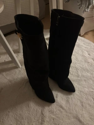 Svarta knähöga stövlar med klack - Snygga svarta mocha stövlar med spetsig tå och bred klack. Dragkedja på insidan och dekorativt guldfärgat spänne upptill. Använda 2 gånger och nypris 600kr.