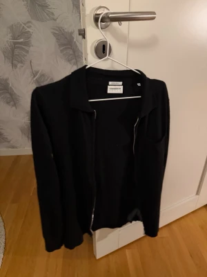 Svart kofta från Lindbergh - Stilren svart kofta från Lindbergh med dragkedja framtill och klassisk krage. Perfekt att slänga på sig över en t-shirt eller skjorta. Enkel och tidlös design som passar till det mesta. Nypris 1200