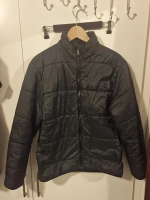 Svart pufferjacka med dragkedja - Stilren svart pufferjacka med quiltad design och hög krage. Jackan har hel dragkedja framtill och långa ärmar. Perfekt för kyliga dagar när du vill hålla värmen och ändå se snygg ut.