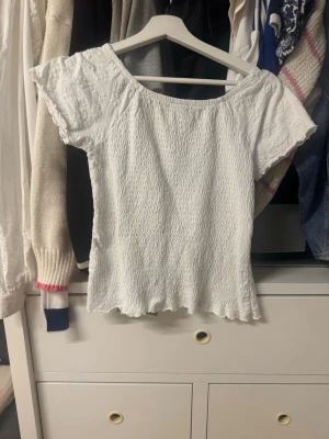 Vit smockad topp med spetsdetaljer - Söt vit topp med smockad kropp och korta ärmar. Ärmarna har fina spetsdetaljer och toppen har en lätt vågig kant nedtill. Perfekt att styla med jeans eller kjol för en fräsch look.