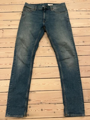 Blå jeans från Tiger of Sweden - Snygga blå jeans från Tiger of Sweden med klassisk femficksmodell och brunt läderpatch bak i midjan. Jeansen har raka ben och normal passform, tillverkade i mjukt bomullsdenim med subtila slitningar framtill för en avslappnad look. Dom är dessutom knappt använda och i riktigt fint skick