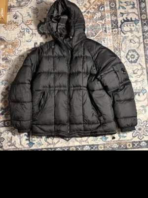 Svart pufferjacka med huva - Säljer en svart pufferjacka med huva och ikonisk linsdetalj på ärmen. Jackan har quiltad design, dragkedja framtill och två sidofickor. Perfekt för kalla dagar och riktigt snygg streetstil. Köpt på Vinted! Så osäker på om den är äkta därav priset!