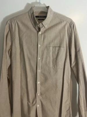 Beige skjorta från Tiger of Sweden  - Stilren beige skjorta från Tiger of Sweden med klassisk button-down krage och en bröstficka. Skjortan har långa ärmar och vita knappar framtill. Perfekt för en clean och enkel look. Materialet känns mjukt och bekvämt.