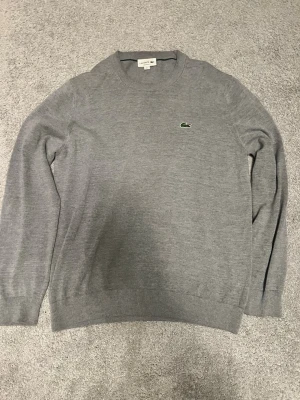 Grå stickad tröja från Lacoste - Säljer en stilren grå stickad tröja från Lacoste med rund halsringning och den klassiska gröna krokodilloggan broderad på bröstet. Tröjan har ribbade muddar vid ärmslut och nederkant. Perfekt till lager på lager och enkel att matcha.
