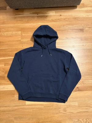 Mörkblå hoodie från RYVLS - En stilren mörkblå hoodie från RYVLS med klassisk känguruficka och justerbar huva med dragsko. Tillverkad i mjukt bomullsmaterial som känns skönt mot huden. Har en liten fläck vid höger axeln som går bort i tvätten.