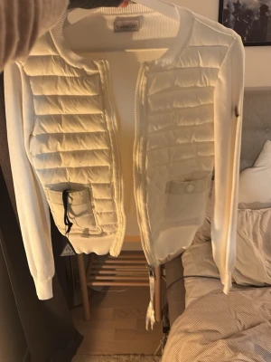 Vit quiltad cardigan jacka Moncler - Stilren vit cardigan jacka från Moncler med quiltade partier framtill och stickade ärmar och rygg. Jackan har dragkedja, rund halsringning och praktiska fickor med knapp och snörning. Perfekt för lager-på-lager under kyliga dagar.