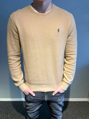 Ralph Lauren tröja  - Snygg beige stickad tröja från Polo Ralph Lauren med rund halsringning och diskret broderad logga på bröstet. Tröjan har en klassisk passform. Tillverkad i mjukt material som känns skönt mot huden. Storlek M.