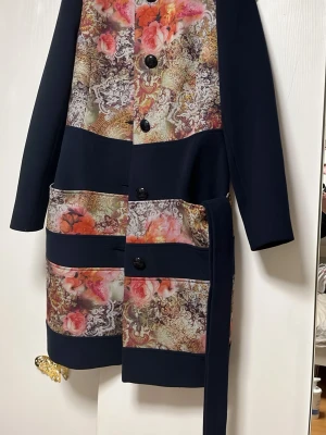 Mönstrad kappa från Ricco Collection - Unik kappa från Ricco Collection med stora svarta knappar och bälte i midjan. Kappan har blommigt mönster i orange, rosa och beige toner samt svarta partier. Långärmad och rak modell, perfekt för dig som vill sticka ut. Mörkblå med mönster i superkvalitet.