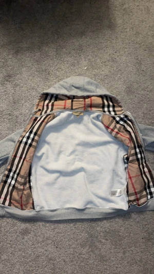 Grå hoodie från Burberry med dragkedja - Snygg grå hoodie från Burberry med klassiskt rutigt foder i huvan och insidan. Dragkedja framtill, två fickor och diskret Burberry-logga på bröstet. Tillverkad i mjuk bomullsblandning, perfekt för en chill och stilren look.