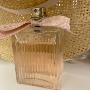 Chloé Eau de Parfum med rosa rosett - Elegant parfym från Chloé i en rektangulär glasflaska med räfflad yta och ljusrosa doftvätska. Flaskan pryds av en ljusrosa tygrosett vid halsen och har en metallfärgad spraykork. Perfekt för dig som gillar feminina och fräscha dofter.