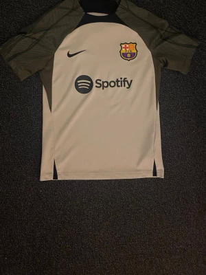 Barcelona tränings t-shirt Nike beige - Snygg beige Barcelona tränings t-shirt från Nike med svarta detaljer och klubbmärke på bröstet. Spotify-logga tryckt på framsidan. Perfekt för fotbollsträning eller att bära till vardags. Korta ärmar och sportig passform.